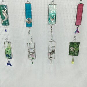 May include: A collection of eight stained glass ornaments featuring various sea life designs. The ornaments are suspended from silver hooks and have dangling charms. The designs include a mermaid, a dolphin, a seahorse, a starfish, a seashell, and a wave.