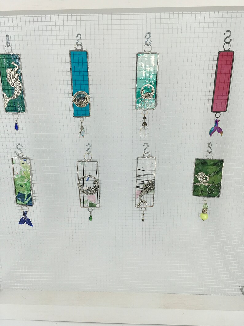 May include: A collection of eight stained glass ornaments featuring various sea life designs. The ornaments are suspended from silver hooks and have dangling charms. The designs include a mermaid, a dolphin, a seahorse, a starfish, a seashell, and a wave.