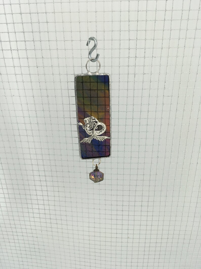 May include: A stained glass suncatcher featuring a mermaid design with a purple iridescent crystal hanging below. The suncatcher is framed in silver and has a dark background with shades of purple, blue, and green.