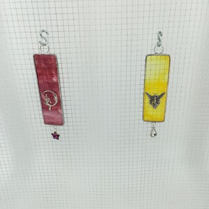 May include: Two stained glass suncatchers hanging from a wire mesh. The left suncatcher is pink and features a fairy with a crescent moon. The right suncatcher is yellow and features a butterfly.