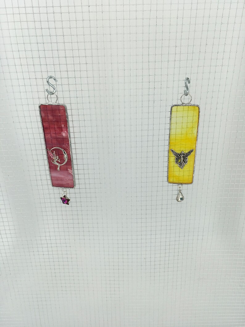 May include: Two stained glass suncatchers hanging from a wire mesh. The left suncatcher is pink and features a fairy with a crescent moon. The right suncatcher is yellow and features a butterfly.