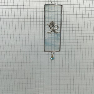May include: A blue and white stained glass suncatcher with a silver mermaid charm hanging from it. The suncatcher is rectangular and has a silver chain attached to the top.