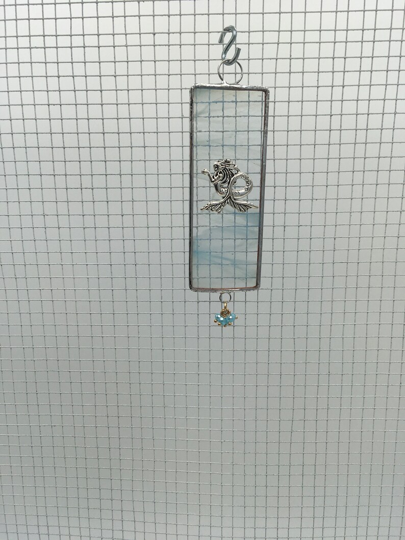 May include: A blue and white stained glass suncatcher with a silver mermaid charm hanging from it. The suncatcher is rectangular and has a silver chain attached to the top.