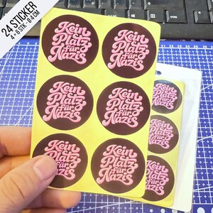 Könnte beinhalten: Ein Bogen mit runden Aufklebern, die den Text Kein Platz für Nazis in rosa Schreibschrift auf dunkelbraunem Hintergrund zeigen. Der Bogen enthält 24 Aufkleber mit einem Durchmesser von jeweils 4 cm, die in einem Raster angeordnet sind.