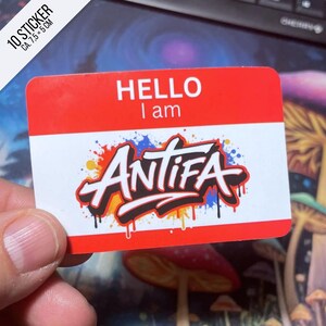 Könnte beinhalten: Ein rechteckiger Aufkleber mit rotem Oberteil und weißem Unterteil. Der Text "HELLO I am" ist in Schwarz gedruckt. Das Wort "ANTIFA" ist in einer Graffiti-Schriftart mit bunten Farbspritzern gehalten. Der Aufkleber misst ca. 7,5 x 5 cm.