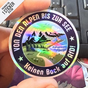 Könnte beinhalten: Ein kreisförmiger holografischer Aufkleber mit 6 cm Durchmesser, der eine Landschaft mit Bergen, einem Weg und einem Leuchtturm zeigt. Der Text lautet "VON DEN ALPEN BIS ZUR SEE" und "Keinen Bock auf AfD!", getrennt durch Sterne.