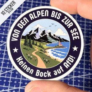 Könnte beinhalten: Ein runder Aufkleber mit einer Landschaftsillustration aus schneebedeckten Bergen, einem geschwungenen Pfad, einem Leuchtturm und blauem Meer. Der dunkelblaue Rand enthält den weißen Text VON DEN ALPEN BIS ZUR SEE und Keinen Bock auf AfD!.