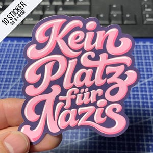 Könnte beinhalten: Ein gestanzter Aufkleber mit dem Text "Kein Platz für Nazis" in einer fetten, rosafarbenen Bubble-Schrift mit dunkelvioletter Umrandung. Der Aufkleber misst ca. 8 x 8 cm, wie auf der Ecke der Verpackung angegeben.