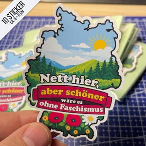 Könnte beinhalten: Ein konturgestanzter Aufkleber in Form der Deutschlandkarte mit einer Landschaft aus gelber Sonne, blauem Himmel, weißen Wolken, grünen Hügeln und Tannen. Der Text lautet: Nett hier, aber schöner wäre es ohne Faschismus, mit roten Mohnblumen.