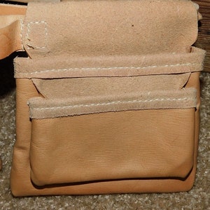 May include: Close-up of a tan leather tool belt pouch with multiple pockets. The pouch is made of smooth leather with suede trim and white stitching. The design is for carrying tools.