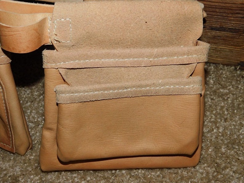 May include: Close-up of a tan leather tool belt pouch with multiple pockets. The pouch is made of smooth leather with suede trim and white stitching. The design is for carrying tools.
