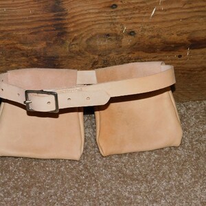 May include: A light tan leather belt with two attached pouches. The belt has a silver-colored buckle and is designed to be worn around the waist. The pouches are rectangular and appear to be empty. The belt is likely for carrying tools or other small items.