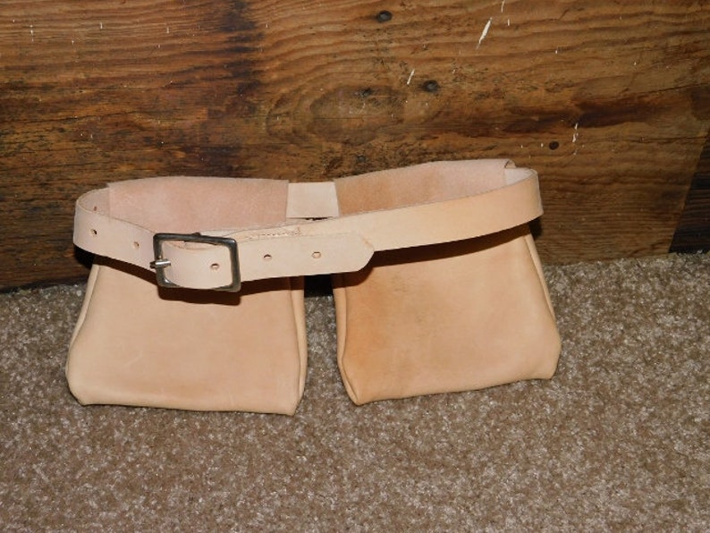 May include: A light tan leather belt with two attached pouches. The belt has a silver-colored buckle and is designed to be worn around the waist. The pouches are rectangular and appear to be empty. The belt is likely for carrying tools or other small items.