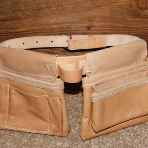 May include: A light brown leather tool belt with multiple pockets and a belt. The belt has several holes for adjustment and a metal buckle. The tool belt is designed to be worn around the waist.