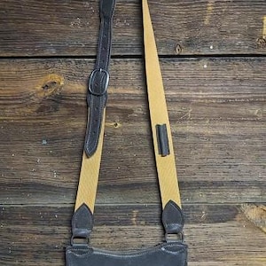 May include: A dark gray suede crossbody bag with a tan strap and dark brown leather accents. The bag has a curved bottom and a flap closure. The strap is adjustable with a buckle. The bag is hanging against a wooden background.