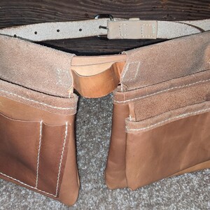 May include: A two-pocket leather tool belt with a light brown belt and buckle. The tool belt has multiple pockets and is stitched with white thread. The leather has a natural, worn appearance.