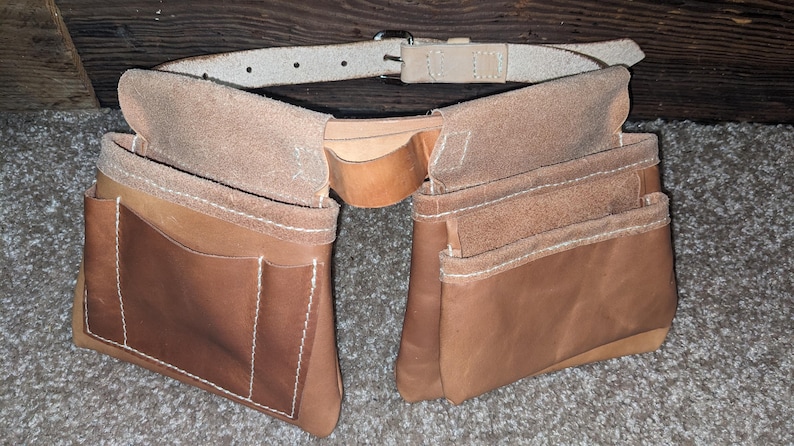 May include: A two-pocket leather tool belt with a light brown belt and buckle. The tool belt has multiple pockets and is stitched with white thread. The leather has a natural, worn appearance.