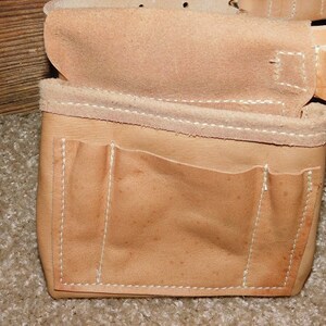 May include: Close-up of a tan leather tool belt with multiple pockets and compartments. The belt features white stitching and is designed for carrying tools. The leather has a natural, worn appearance.