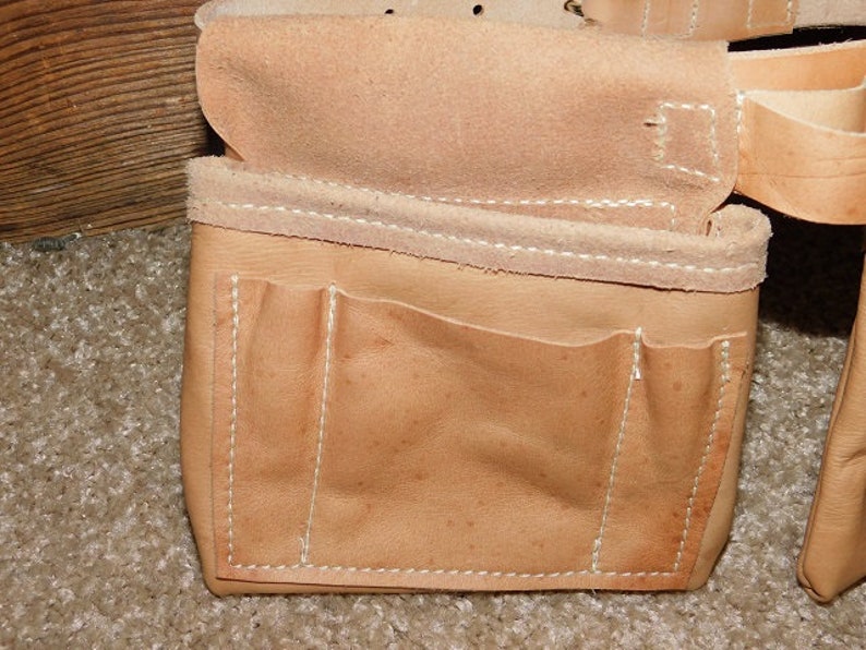 May include: Close-up of a tan leather tool belt with multiple pockets and compartments. The belt features white stitching and is designed for carrying tools. The leather has a natural, worn appearance.