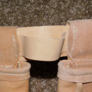 May include: Close-up of a light tan leather belt loop with a matching belt. The belt loop is stitched with white thread. The background is a textured, light brown surface.