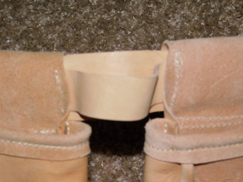 May include: Close-up of a light tan leather belt loop with a matching belt. The belt loop is stitched with white thread. The background is a textured, light brown surface.