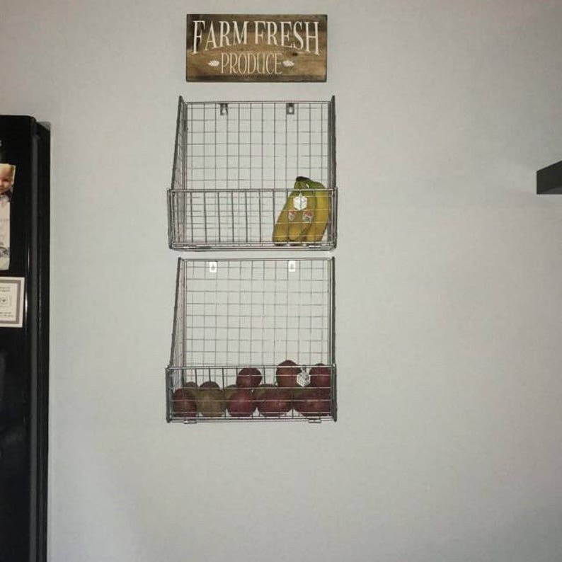 K&ouml;nnte beinhalten: Zwei Drahtk&ouml;rbe an einer Wand montiert, einer &uuml;ber dem anderen. Der obere Korb enth&auml;lt Bananen, der untere Korb enth&auml;lt rote &Auml;pfel. Ein Holzschild &uuml;ber den K&ouml;rben lautet "Farm Fresh Produce".