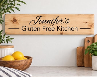 May include: A rectangular wooden sign with a natural light brown wood grain finish. The sign features black text that reads "Jennifer's" in a cursive script above the words "Gluten Free Kitchen" in a sans-serif font, flanked by horizontal lines.