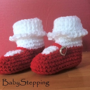 May include: A pair of red and white crocheted baby booties with a white ruffle and a red heart-shaped button. The booties are on a wooden surface.
