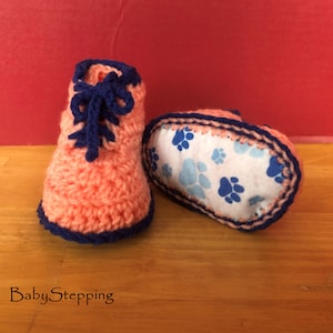 May include: Handmade crochet baby booties in peach with navy blue accents. The booties have navy blue laces and trim. The sole of one bootie is visible, featuring a white fabric with blue paw prints. The text "BabyStepping" is visible.