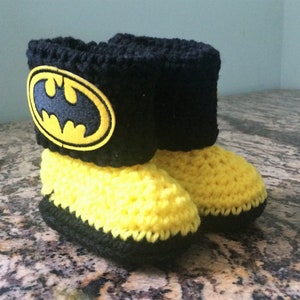 Op de afbeelding: Gele en zwarte gehaakte babyschoentjes met een zwart Batman-logo op de bovenkant.