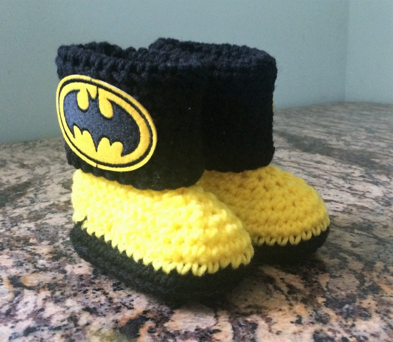 Op de afbeelding: Gele en zwarte gehaakte babyschoentjes met een zwart Batman-logo op de bovenkant.