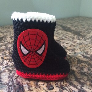 Op de afbeelding: Zwarte en rode gehaakte babyschoentjes met een wit en rood geborduurd Spiderman-gezicht aan de voorkant.