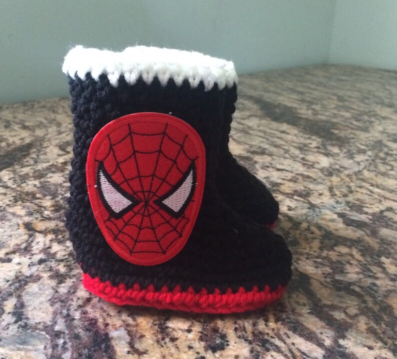 Op de afbeelding: Zwarte en rode gehaakte babyschoentjes met een wit en rood geborduurd Spiderman-gezicht aan de voorkant.