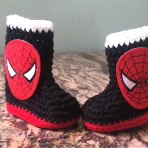 Op de afbeelding: Een paar zwarte en rode gehaakte babyschoentjes met een witte rand en een rode en zwarte Spiderman-applicatie op elke schoen.