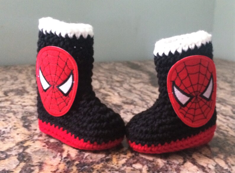 Op de afbeelding: Een paar zwarte en rode gehaakte babyschoentjes met een witte rand en een rode en zwarte Spiderman-applicatie op elke schoen.