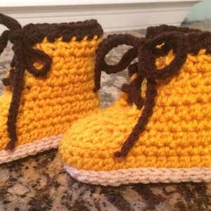 May include: A pair of yellow crocheted baby booties with brown laces. The booties have a white crocheted trim around the bottom.