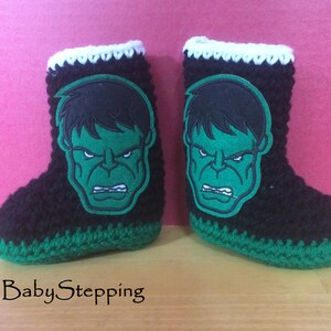 Op de afbeelding: Een paar zwarte, groene en witte gehaakte babyschoentjes met een groene Hulk-gezichtsapplicatie op elke schoen.