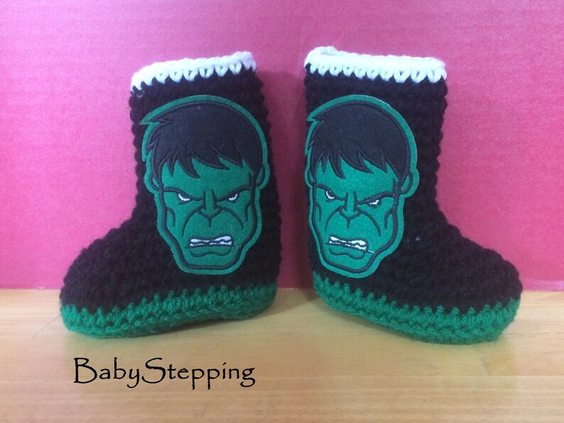Op de afbeelding: Een paar zwarte, groene en witte gehaakte babyschoentjes met een groene Hulk-gezichtsapplicatie op elke schoen.
