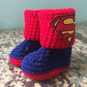 Op de afbeelding: Rode en blauwe gehaakte babyschoentjes met een Superman-logo op de bovenkant van een schoentje.