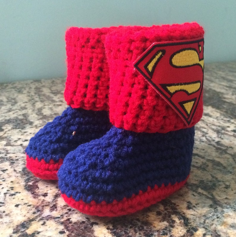 Op de afbeelding: Rode en blauwe gehaakte babyschoentjes met een Superman-logo op de bovenkant van een schoentje.