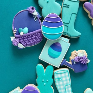 May include: A collection of Easter-themed sugar cookies decorated with purple and blue icing. The cookies are shaped like bunnies, eggs, a watering can, a basket, and rain boots.