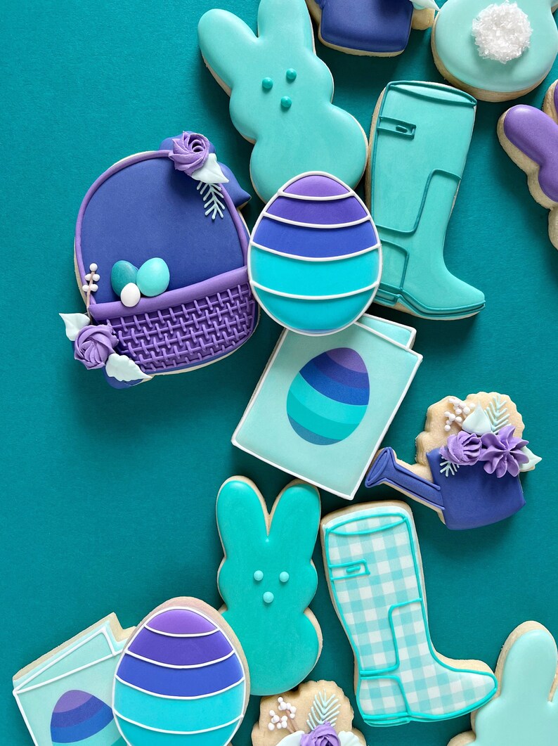 May include: A collection of Easter-themed sugar cookies decorated with purple and blue icing. The cookies are shaped like bunnies, eggs, a watering can, a basket, and rain boots.