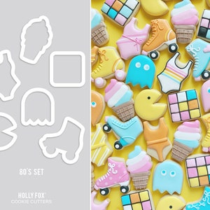 Pode incluir: Um conjunto de cortadores de biscoitos com tema dos anos 80, incluindo um fantasma, um Pac-Man, um patins, um quadrado, um fato de banho, um cone de gelado e um fantasma. Os cortadores de biscoitos são brancos e estão sobre um fundo cinzento. O texto "80's SET" e "HOLLY FOX COOKIE CUTTERS" está abaixo dos cortadores de biscoitos.