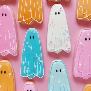 Puede incluir: Una colecci&oacute;n de nueve galletas con forma de fantasma decoradas con diferentes colores y purpurina. Las galletas est&aacute;n dispuestas sobre un fondo rosa.