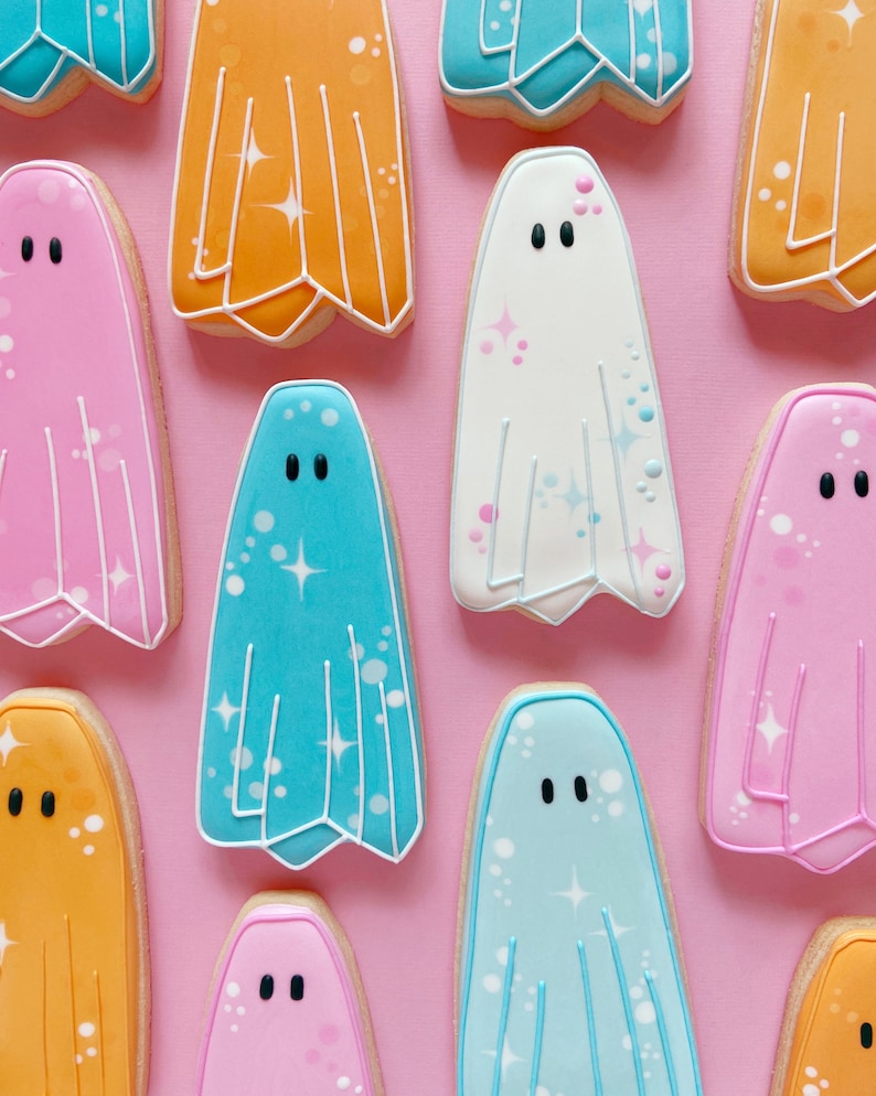 Puede incluir: Una colecci&oacute;n de nueve galletas con forma de fantasma decoradas con diferentes colores y purpurina. Las galletas est&aacute;n dispuestas sobre un fondo rosa.
