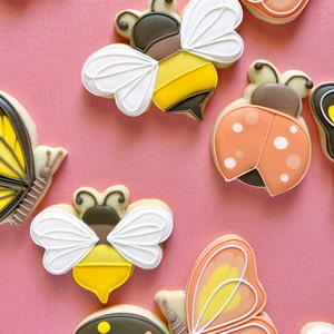 Pode incluir: Uma cole&ccedil;&atilde;o de biscoitos de a&ccedil;&uacute;car coloridos decorados com v&aacute;rios desenhos de insetos, incluindo borboletas, joaninhas e abelhas. Os biscoitos est&atilde;o dispostos sobre um fundo rosa.