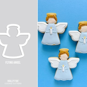May include: A white cookie cutter in the shape of a flying angel next to three angel-shaped cookies. The cookies have light blue dresses, white wings, and gold halos. The cookie cutter has the text "FLYING ANGEL" and "HOLLY FOX COOKIE CUTTERS".