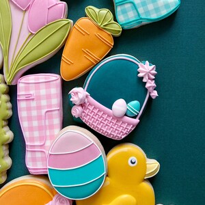 May include: A collection of Easter-themed sugar cookies decorated with colorful icing. The cookies feature a bunny, tulips, carrots, a duck, a basket, and rain boots.