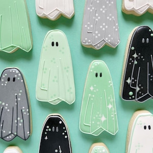 Puede incluir: Una colecci&oacute;n de diez galletas con forma de fantasma decoradas. Las galletas est&aacute;n decoradas con diferentes colores y patrones, incluyendo blanco, gris, negro y verde. Algunas galletas tienen puntos blancos, mientras que otras tienen brillo.