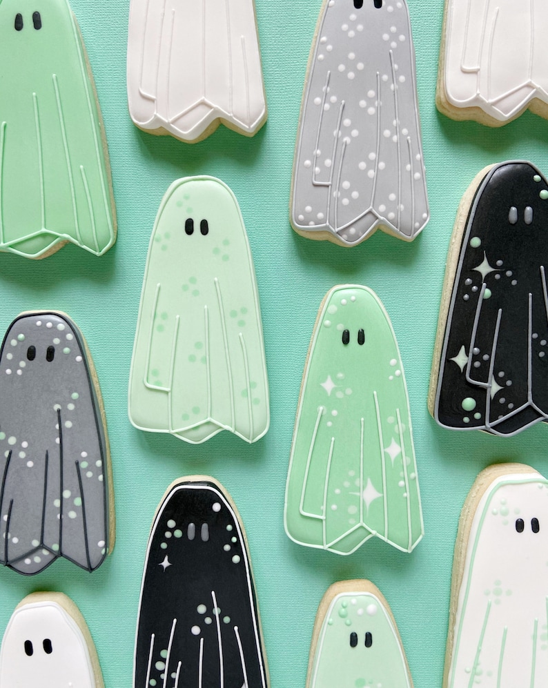 Puede incluir: Una colecci&oacute;n de diez galletas con forma de fantasma decoradas. Las galletas est&aacute;n decoradas con diferentes colores y patrones, incluyendo blanco, gris, negro y verde. Algunas galletas tienen puntos blancos, mientras que otras tienen brillo.